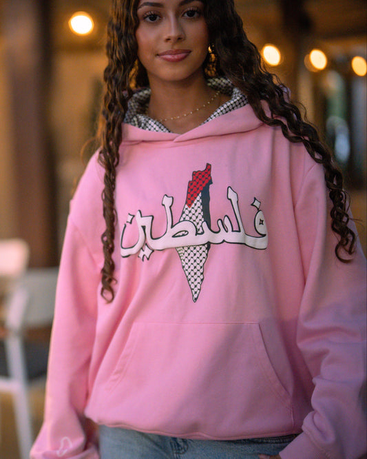Person wearing a pink hoodie with Arabic text in an indoor setting